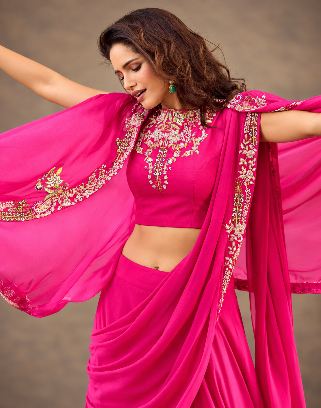 A woman modeling a bright fuchsia pink traditional outfit with an embroidered crop top, draped skirt, and matching sheer dupatta with ornate gold and green embellishments, posed against a neutral brown background.