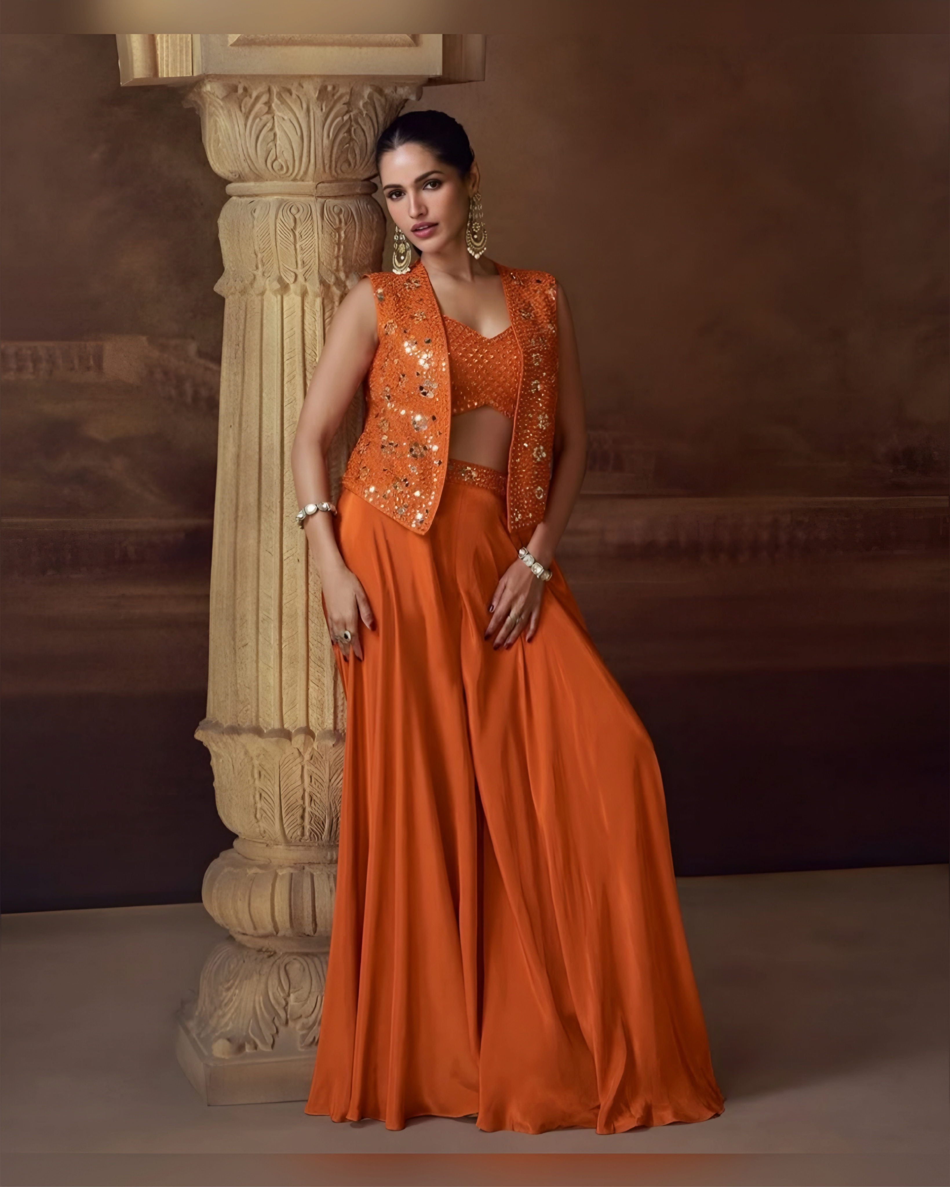 Woman in an orange saree with a matching blouse standing against a decorative wall.