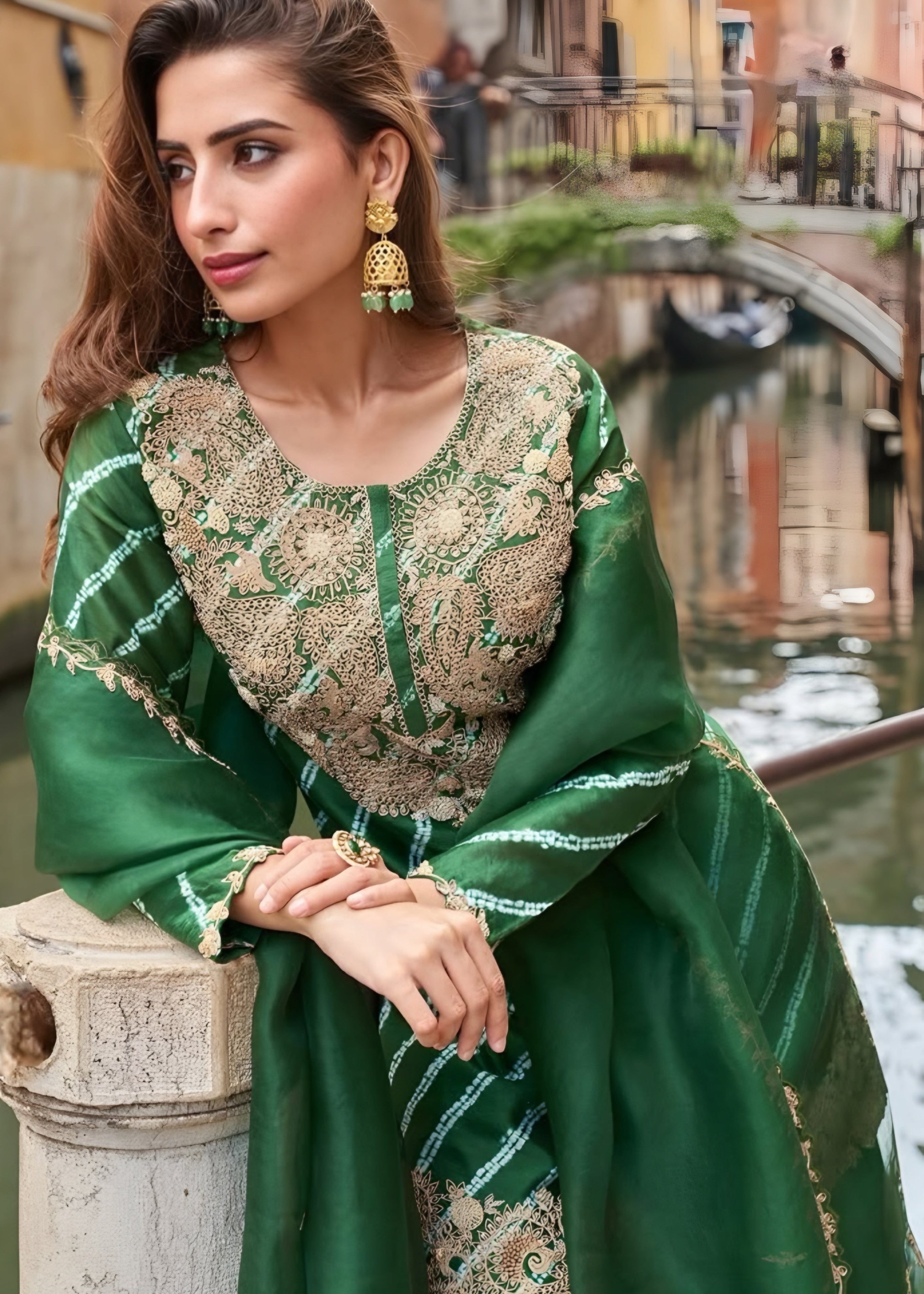 Woman in a green traditional outfit with intricate patterns, standing by a canal with buildings in the background.