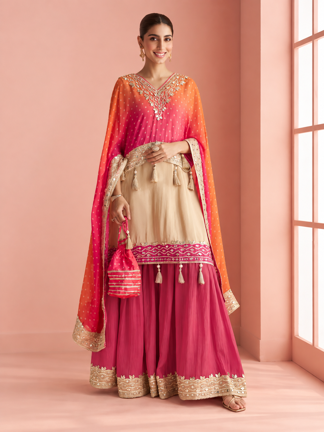 A woman models a traditional Indian outfit consisting of a beige kurta with gold embroidery, a bright pink flared skirt with gold border, and a pink-to-orange ombre dupatta with gold embroidery, set against a pink studio background.