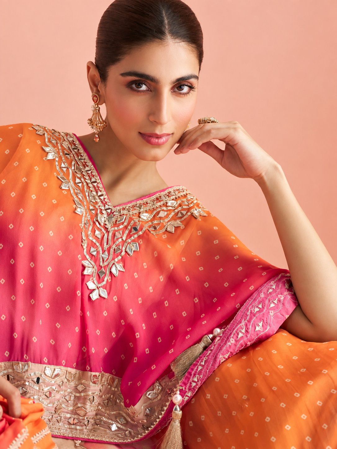 A woman wearing a vibrant pink and orange embroidered traditional sari with metallic silver mirror work and gold detailing, posed against a solid peach-pink background.