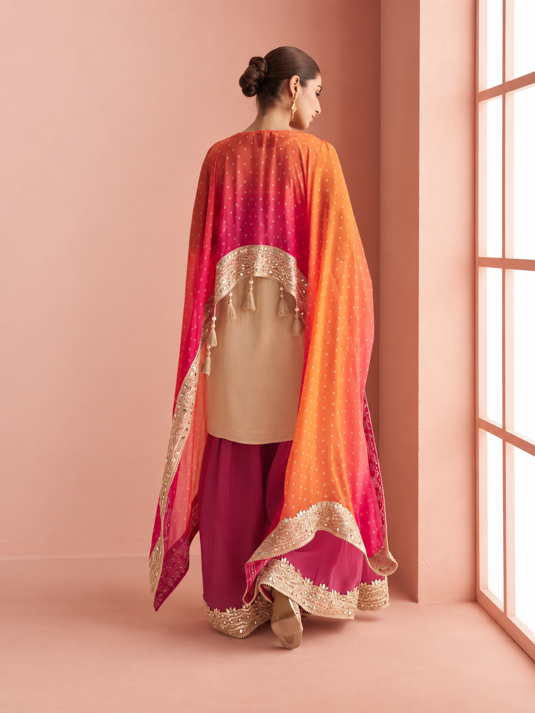 A woman stands with her back to the camera wearing a traditional outfit consisting of a beige kurta, magenta wide-leg pants, and a vibrant orange-to-pink gradient dupatta with gold borders, set against a warm pink studio background.