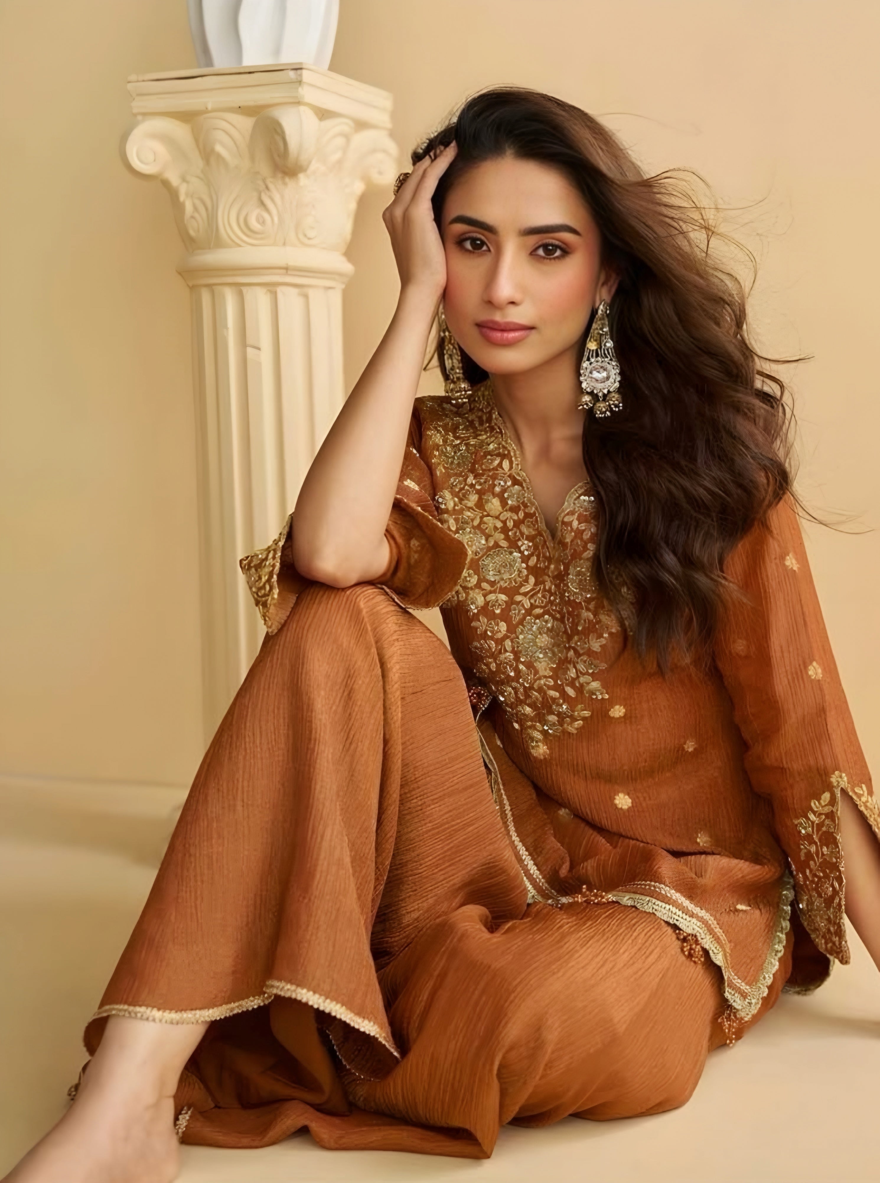 Woman in a traditional brown outfit with gold embroidery sitting against a beige wall.
