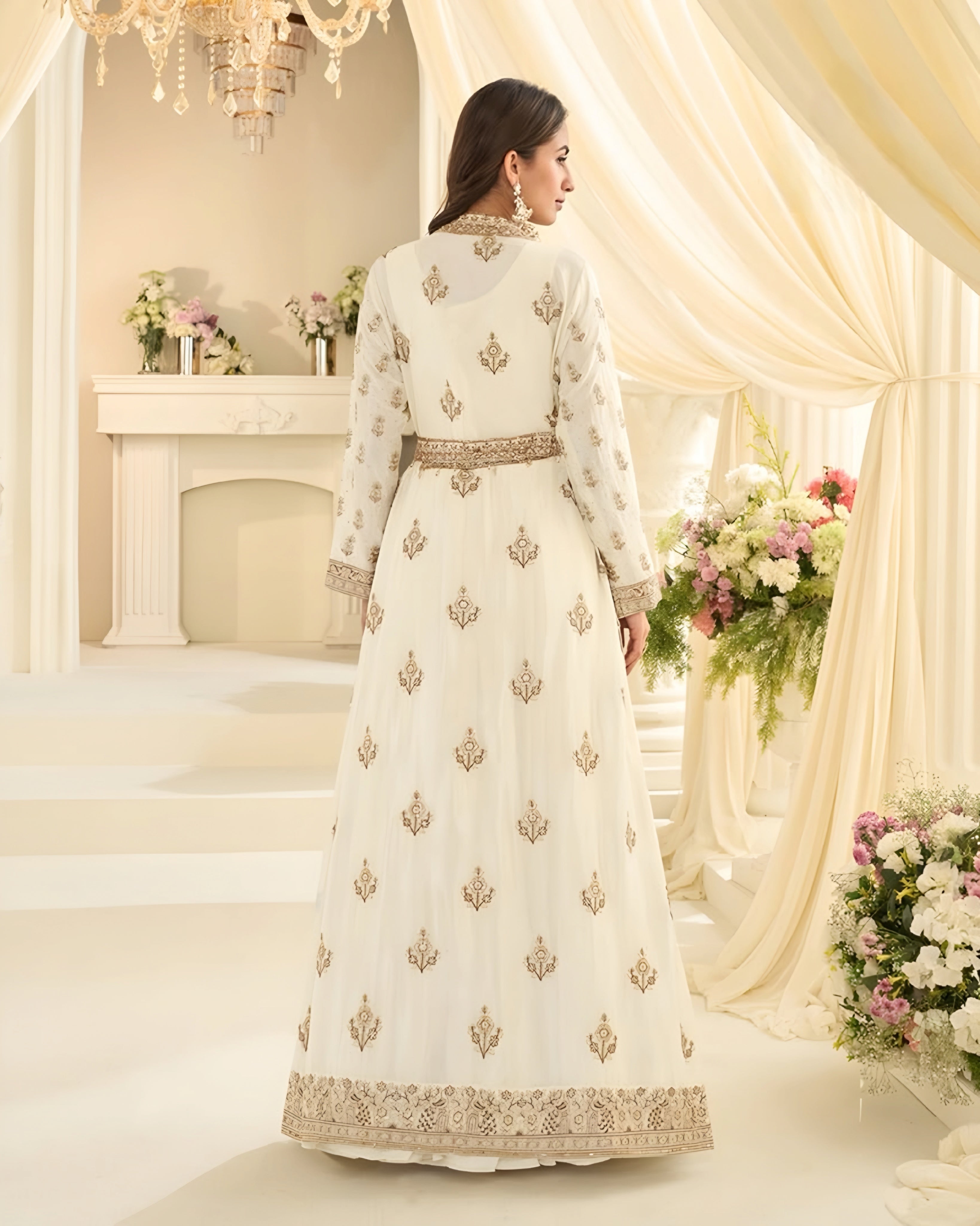 Woman in a white embroidered dress standing in a decorated room with flowers and a chandelier.