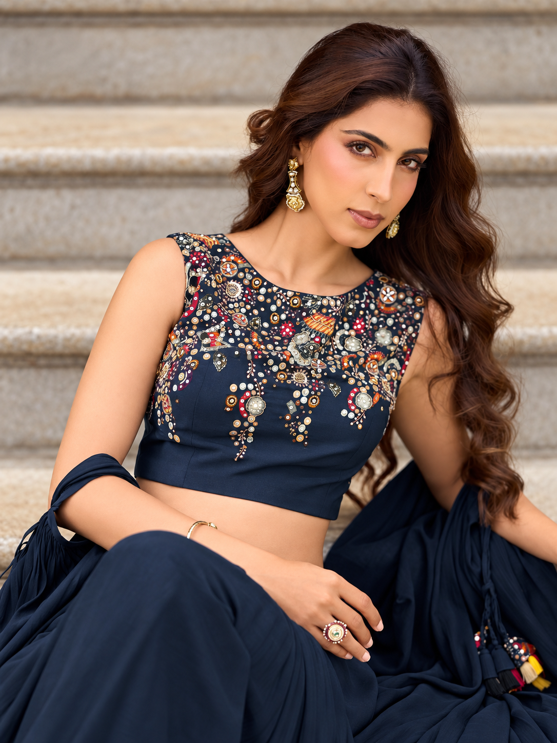 A woman seated on stone steps wearing a navy blue embellished sleeveless top with colorful beadwork and a matching navy skirt or drape.