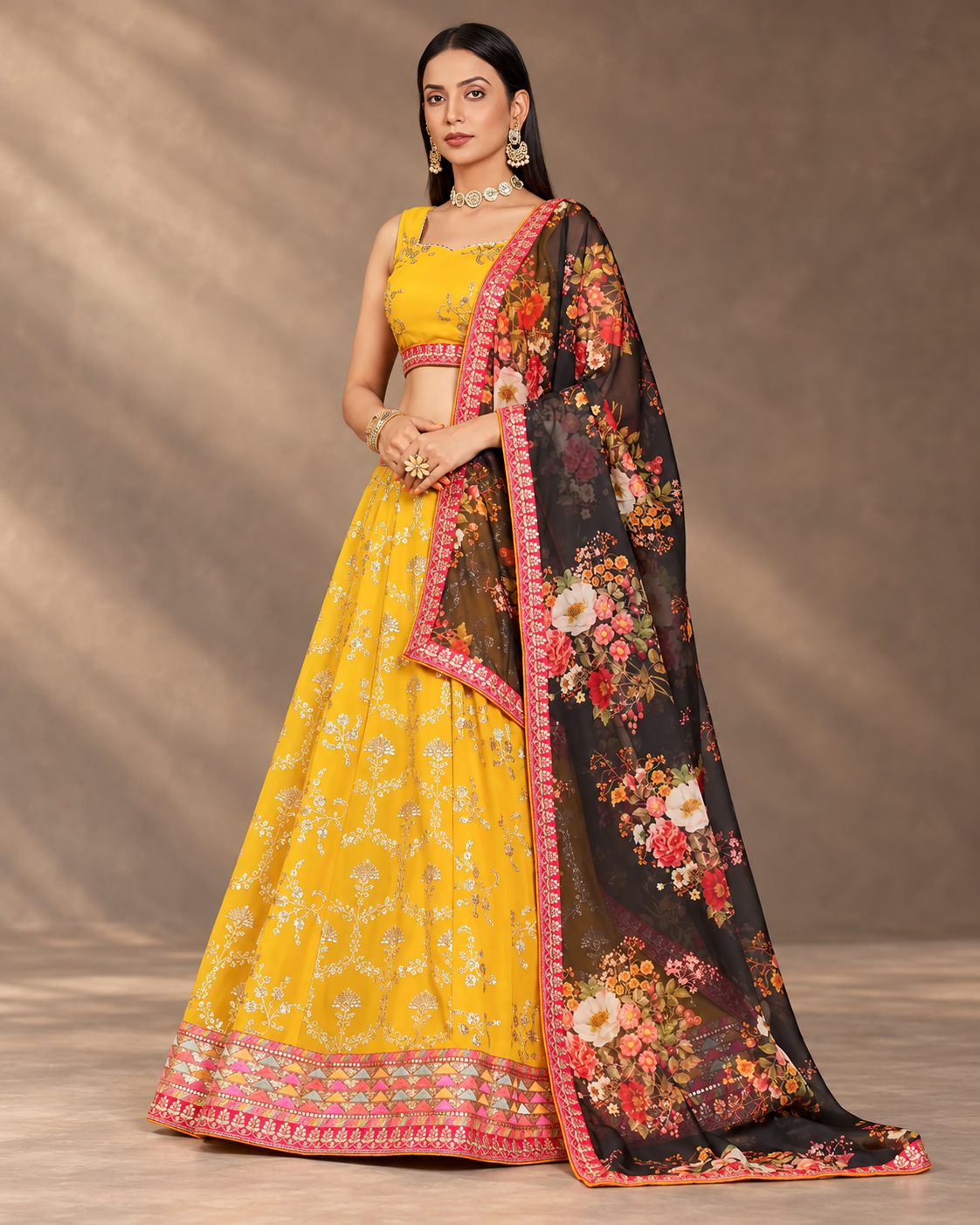 A woman models a traditional Indian lehenga choli set with a floral dupatta. The outfit features a bright yellow skirt and blouse with intricate silver embroidery, paired with a black dupatta decorated with a multicolor floral print and pink borders.