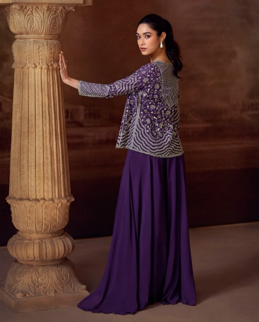 Woman in a purple and silver patterned top and long purple skirt standing next to a stone column.