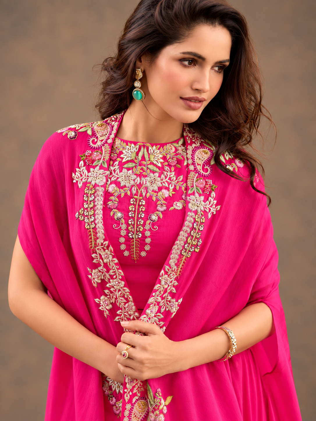 A woman wearing a bright pink traditional outfit with ornate gold and silver embroidery, a matching pink shawl draped over her shoulders, and green teardrop earrings, set against a neutral brown background.