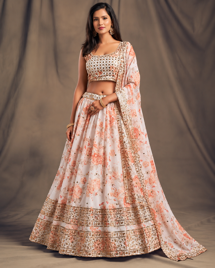 A woman models a traditional Indian lehenga choli set with a dupatta. The outfit is light pink with peach floral prints and detailed gold embroidery, set against a neutral brown studio backdrop.