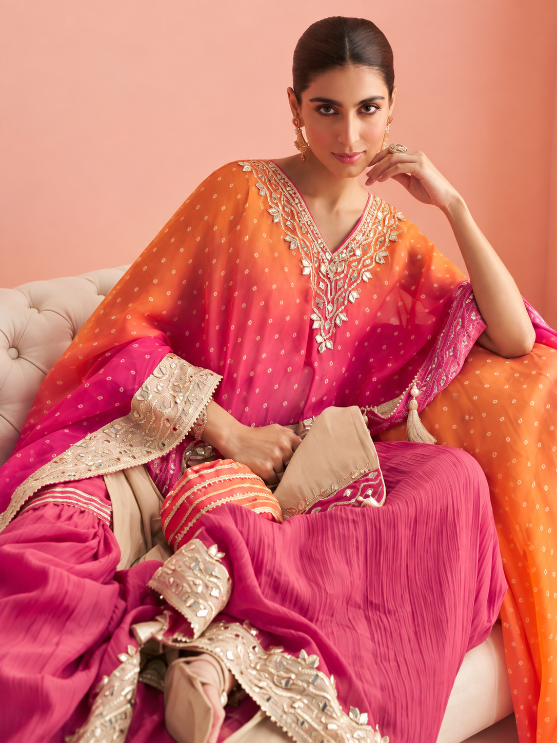 A woman seated on a cream tufted sofa wearing a traditional South Asian outfit with a bright pink and orange draped top and skirt, decorated with ornate gold embroidery and a matching dupatta.