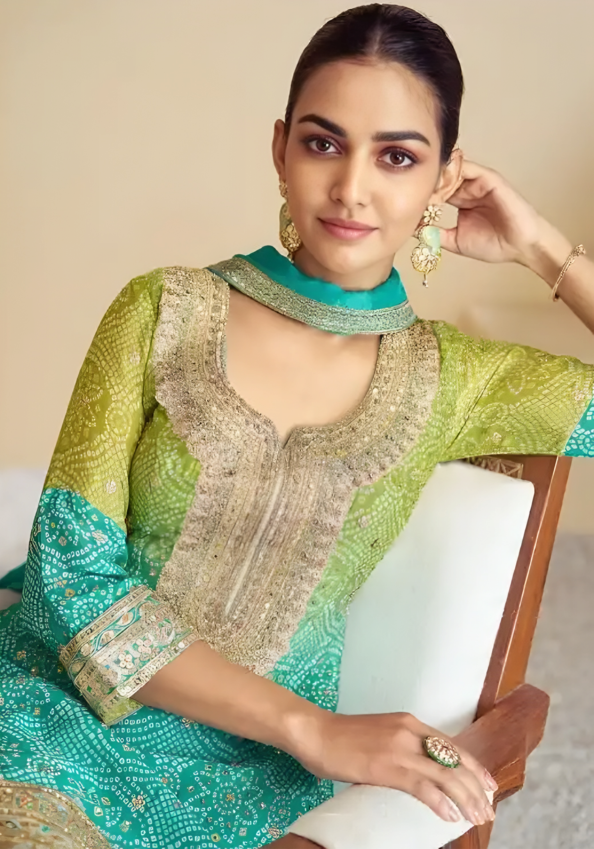 Woman wearing a green and teal traditional outfit with intricate patterns, sitting on a chair.