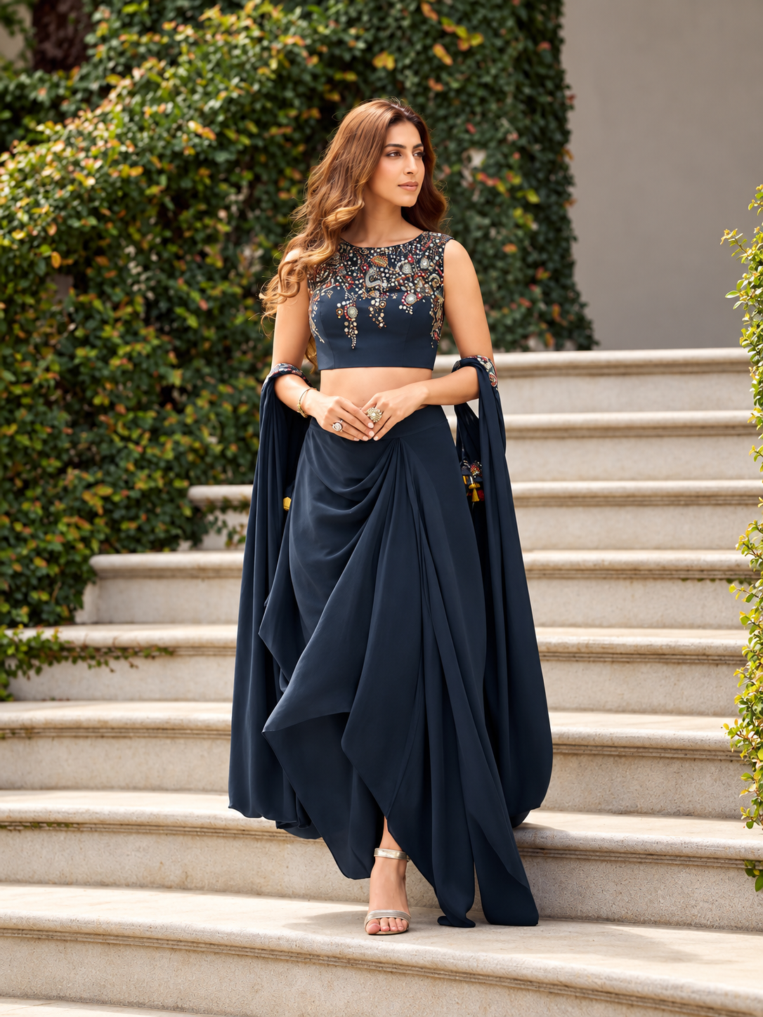 A woman stands on outdoor stone steps wearing a navy blue traditional outfit with a heavily embroidered sleeveless blouse and a draped skirt or dupatta. Greenery and a neutral wall are visible in the background.